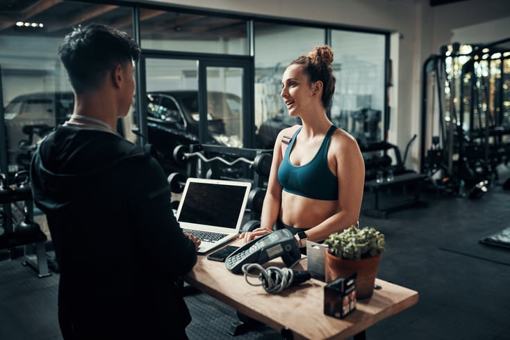 A gym owner talks to one of their gym clients at the front desk of their gym or studio.