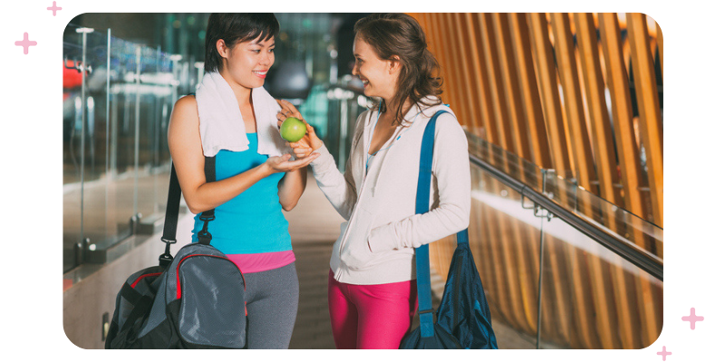 Two women in workout gear. One woman is giving the other woman an apple.