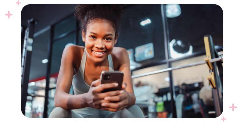 Girl in workout clothes holding phone at the gym, smiling at the camera.