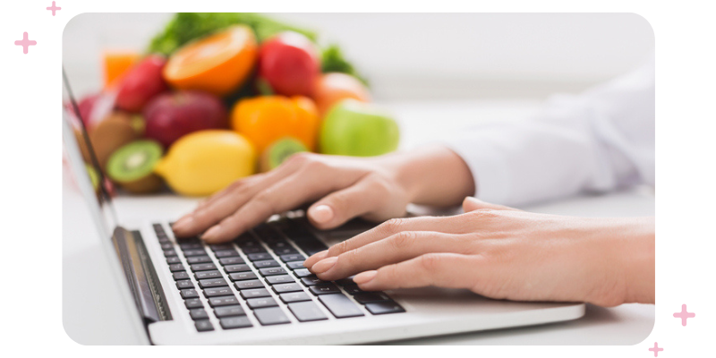 A woman typing on a laptop with a bowl of fruit in the background.