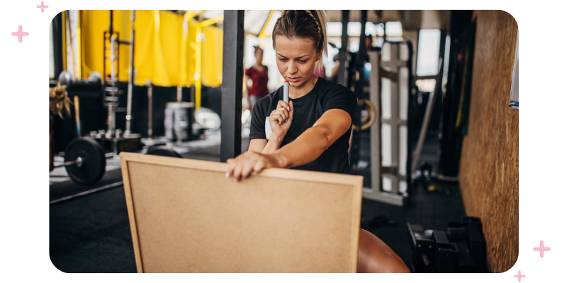 A woman at the gym holding a whiteboard and thinking about what she's going to write.